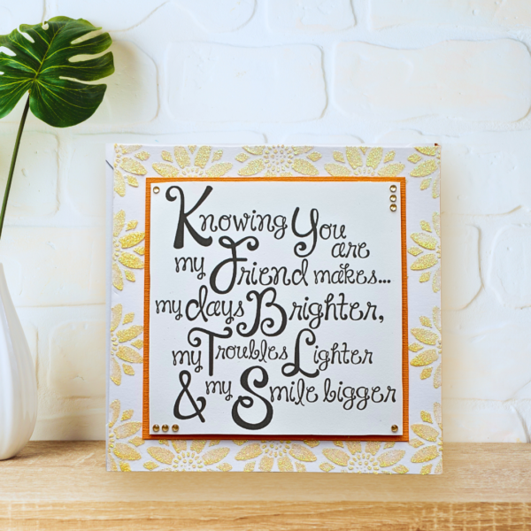 Square card with glittered yelloe flowers and double layered friendship quote displayed on a wooden shelf in front of a white painted brick wall