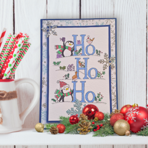 Cute multi layered Christmas card with fun loving penguins and festive Ho Ho Ho displayed on a white shelf in front of a white panelled wall with Christmas decorations.