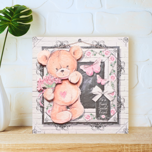 A cure teddy bear with a bunch of pink flowers stood in front of a decorative blackboard with friendship message displayed on a wooden shelf in front of a white brick wall. One of our Teddy Bear Bear Hugs collection.