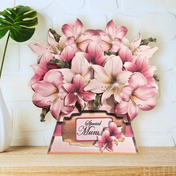 A fully shaped, multi layered amaryllis bouquet card is shown displayed on a wooden shelf in front of a white wall.