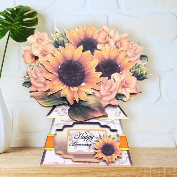 A fully shaped, multi layered sunflower bouquet card is shown displayed on a wooden shelf in front of a white wall.