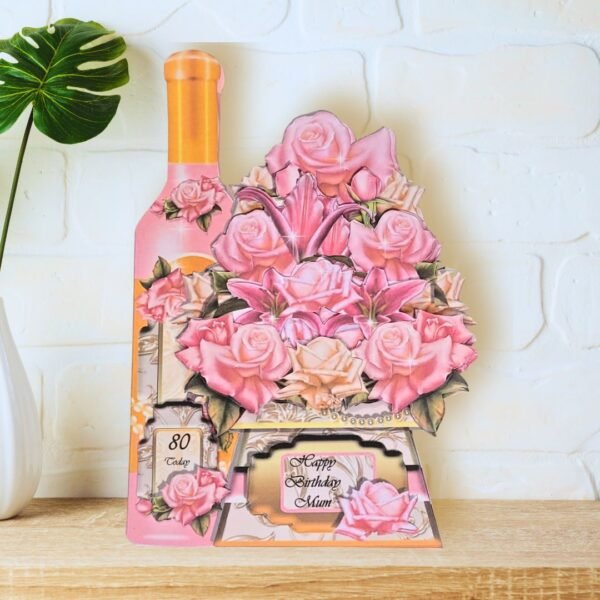 A fully shaped, multi layered rose bouquet and bottle of wine card is shown displayed on a wooden shelf in front of a white wall.