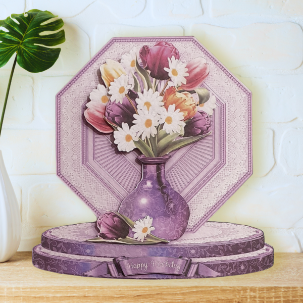 A fully shaped, multi layered dainty tulip & daisy bud vase card is shown displayed on a wooden shelf in front of a white wall.