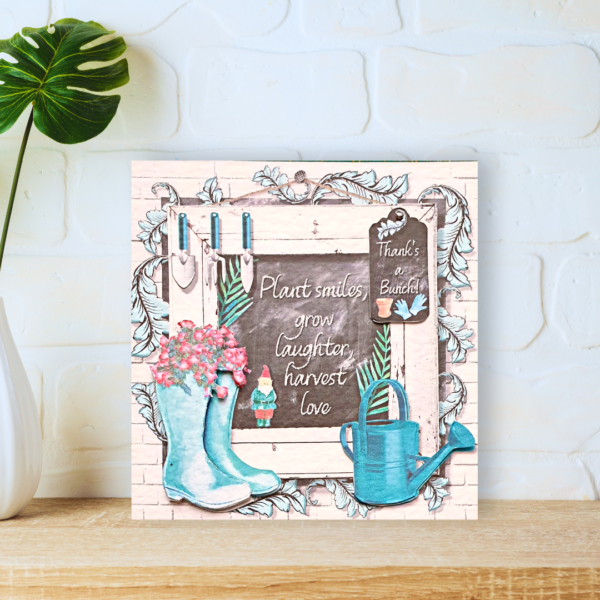 Green Potting Shed Wellies and watering can in front of a blackboard card displayed on a a wooden shelf in front of a white wall.