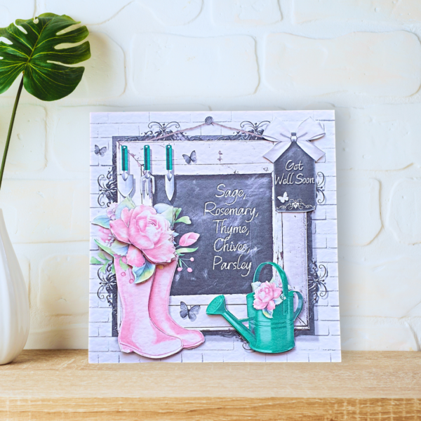 Pink Summerhouse Wellies with pink roses and watering can in front of a blackboard card displayed on a a wooden shelf in front of a white wall.