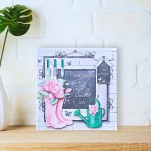 Pink Summerhouse Wellies with pink roses and watering can in front of a blackboard card displayed on a a wooden shelf in front of a white wall.