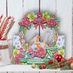 A white shelf in front of white panelled wall with a willow wreath, red Christmas roses and 2 robins.