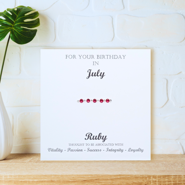 Ruby July Birthstone card displayed on a wooden shelf in front of a white wall.