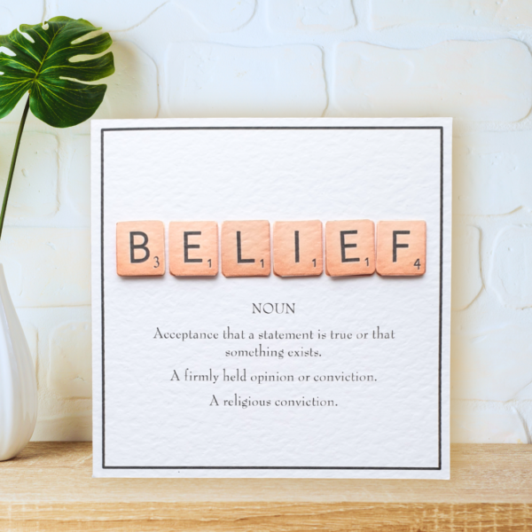 Belief in Scrabble Tiles with printed definition and narrow border, displayed on a wooden shelf in front of a white wall