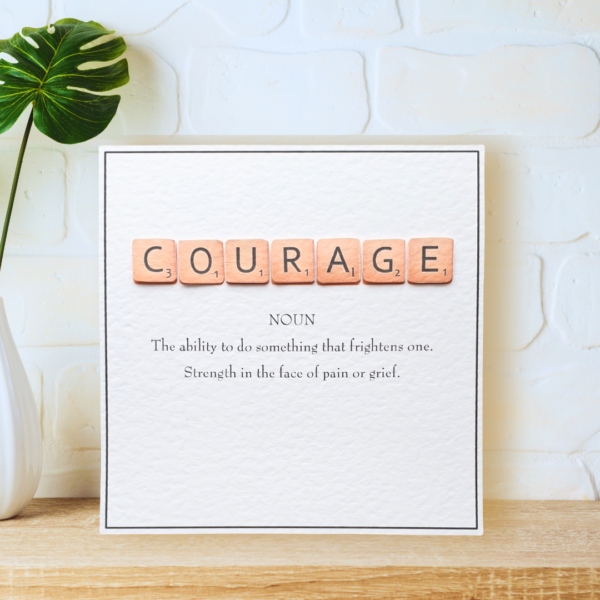 Courage in Scrabble Tiles with printed definition and narrow border, displayed on a wooden shelf in front of a white wall