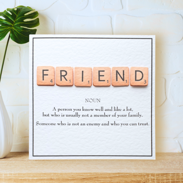 Friend in Scrabble Tiles with printed definition and narrow border, displayed on a wooden shelf in front of a white wall