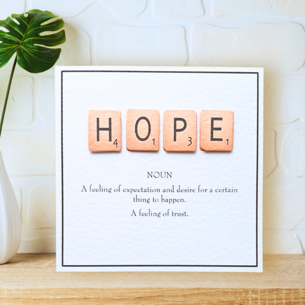 Hope in Scrabble Tiles with printed definition and narrow border, displayed on a wooden shelf in front of a white wall