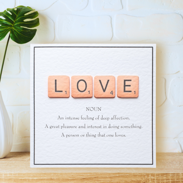 Love in Scrabble Tiles with printed definition and narrow border, displayed on a wooden shelf in front of a white wall