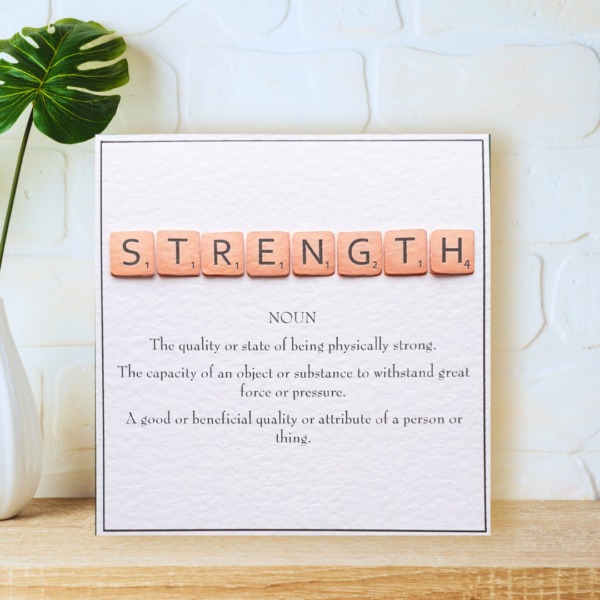 Strength in Scrabble Tiles with printed definition and narrow border, displayed on a wooden shelf in front of a white wall