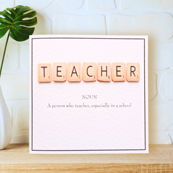 Teacher in Scrabble Tiles with printed definition and narrow border, displayed on a wooden shelf in front of a white wall