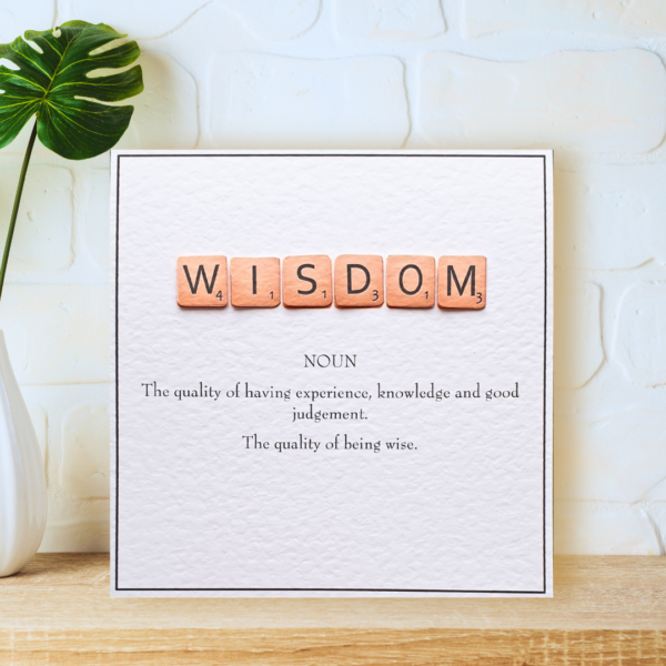 Wisdom in Scrabble Tiles with printed definition and narrow border, displayed on a wooden shelf in front of a white wall