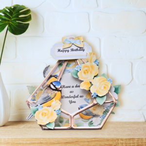 Terrarium shaped card with yellow roses and bluetit displayed on a wooden shelf in front of a white wall.